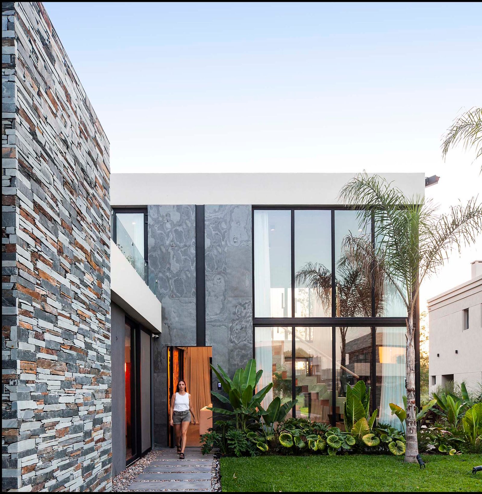 Fachada moderna con muro de piedra, vidrio y vegetación — proyecto CASA T199, por A'PRIMA - Arquitectura Sustentable, en Los Robles 5901