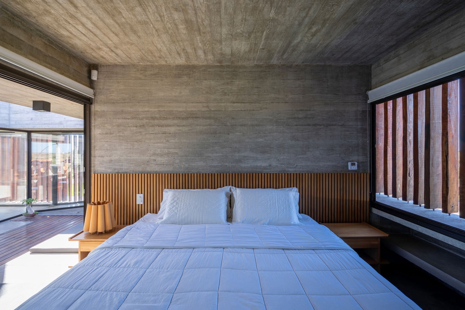 Quarto moderno com paredes de concreto aparente e parede de ripas de madeira — projeto Casa Médanos, por Besonías Almeida arquitectos, em JWRC+MQ