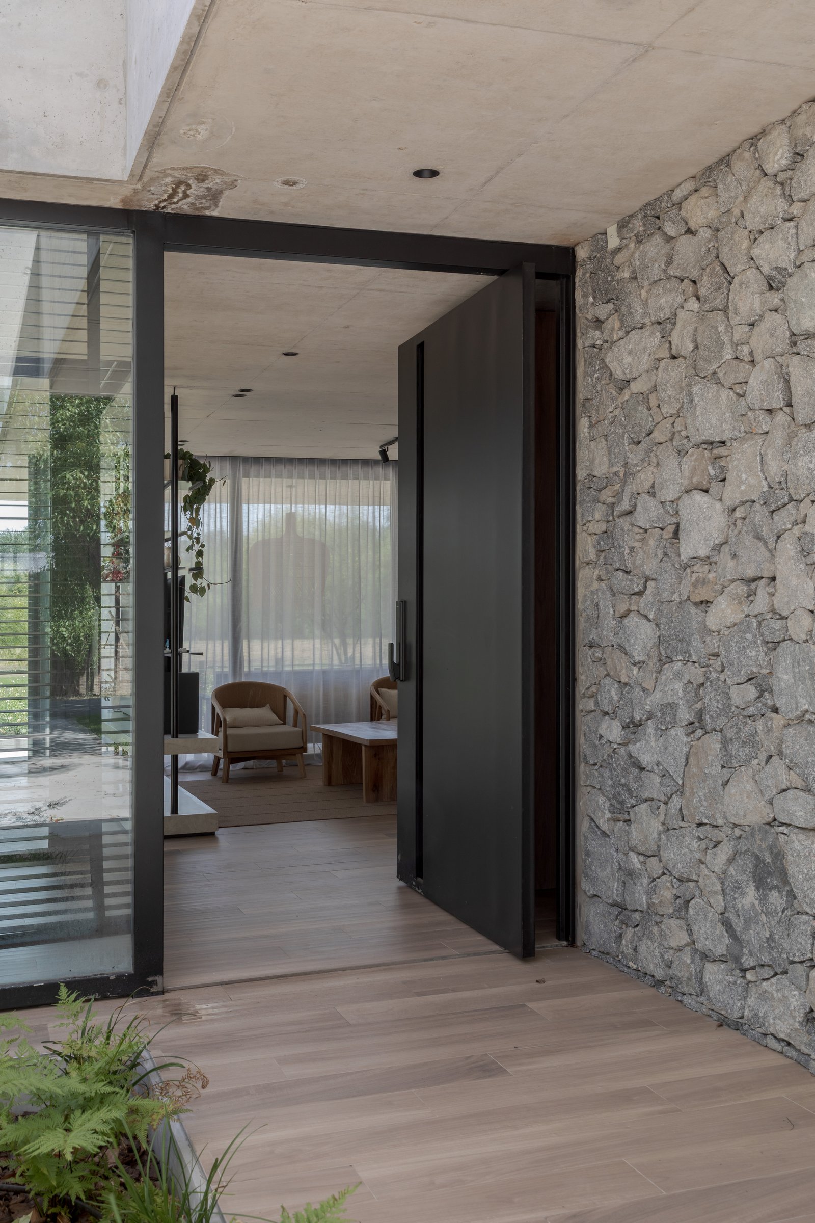 Puerta pivotante de metal negro y muro de piedra en acceso residencial — proyecto Casa PLANALTO, por Grupo CRBN, en Av. Pres. Perón 10298