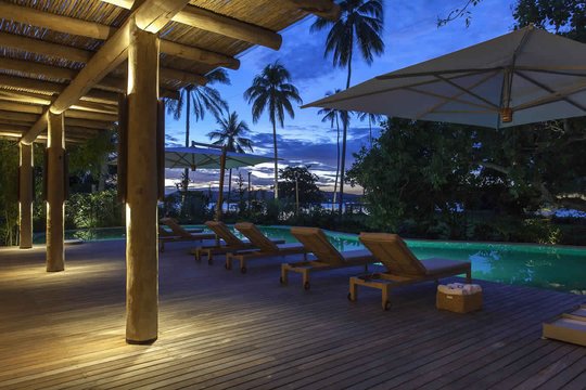 Deck de madera con pileta iluminada al atardecer, palmeras y pergola — proyecto Externas, por Xico Diniz