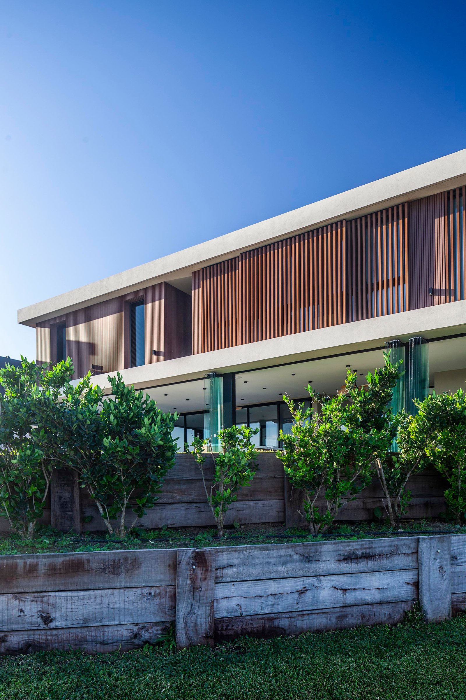 Fachada moderna con revestimiento de madera y muros de contención — proyecto CASA C313, por A'PRIMA - Arquitectura Sustentable, en Provincia de Buenos Aires