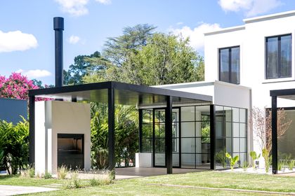 Fachada moderna con pérgola de metal negro y ventanales en jardín soleado — proyecto Bosque Chico, por Estudio Arq. María Sioli, en Garin