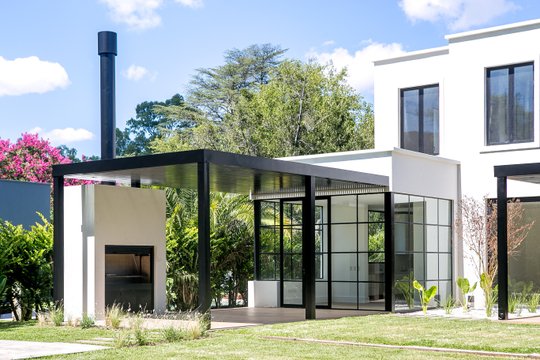 Fachada moderna con pérgola de metal negro y ventanales en jardín soleado — proyecto Bosque Chico, por Estudio Arq. María Sioli, en Garin