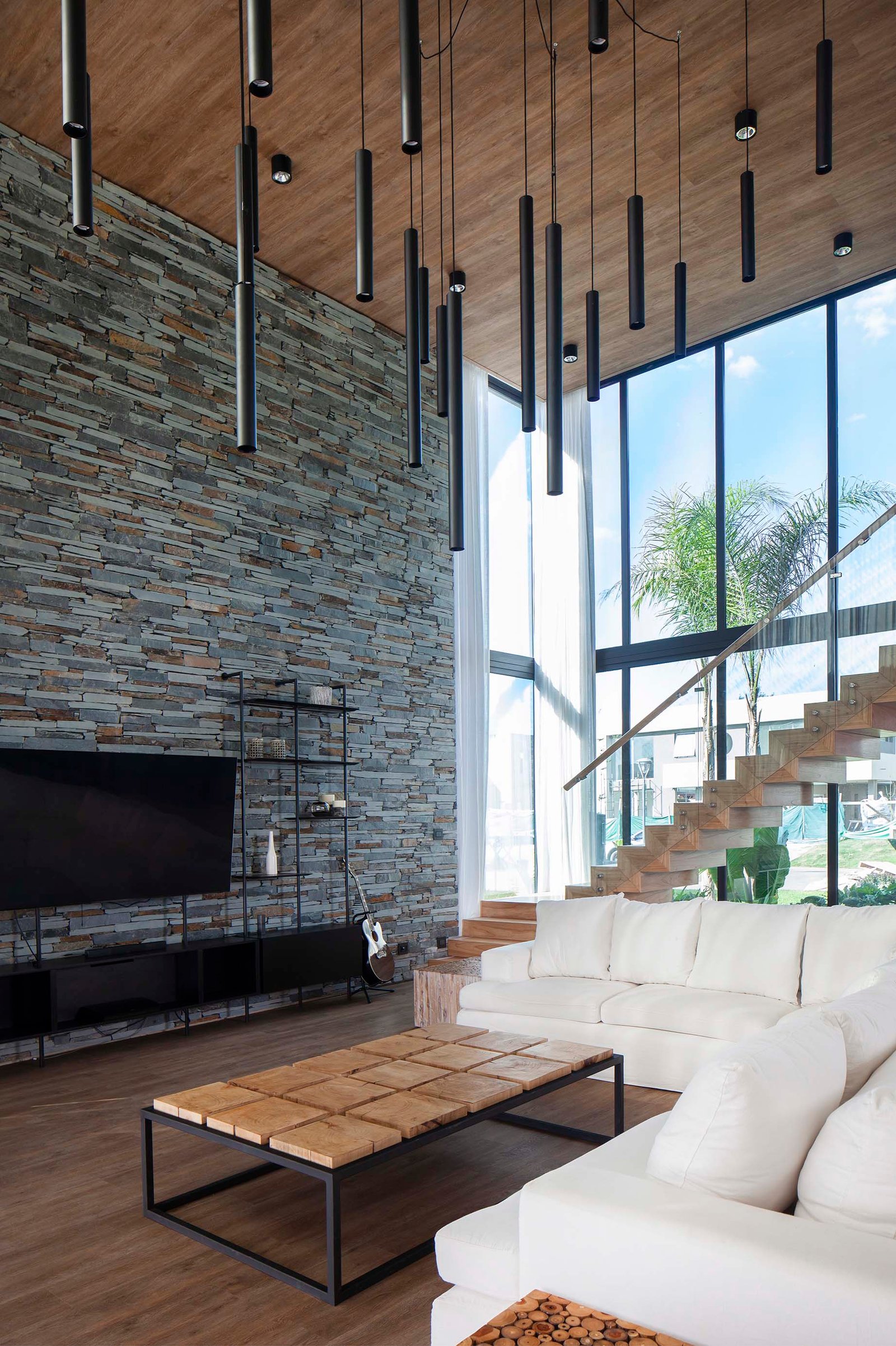 Living moderno con pared de piedra, ventanales y escalera flotante — proyecto CASA T199, por A'PRIMA - Arquitectura Sustentable, en Los Robles 5901