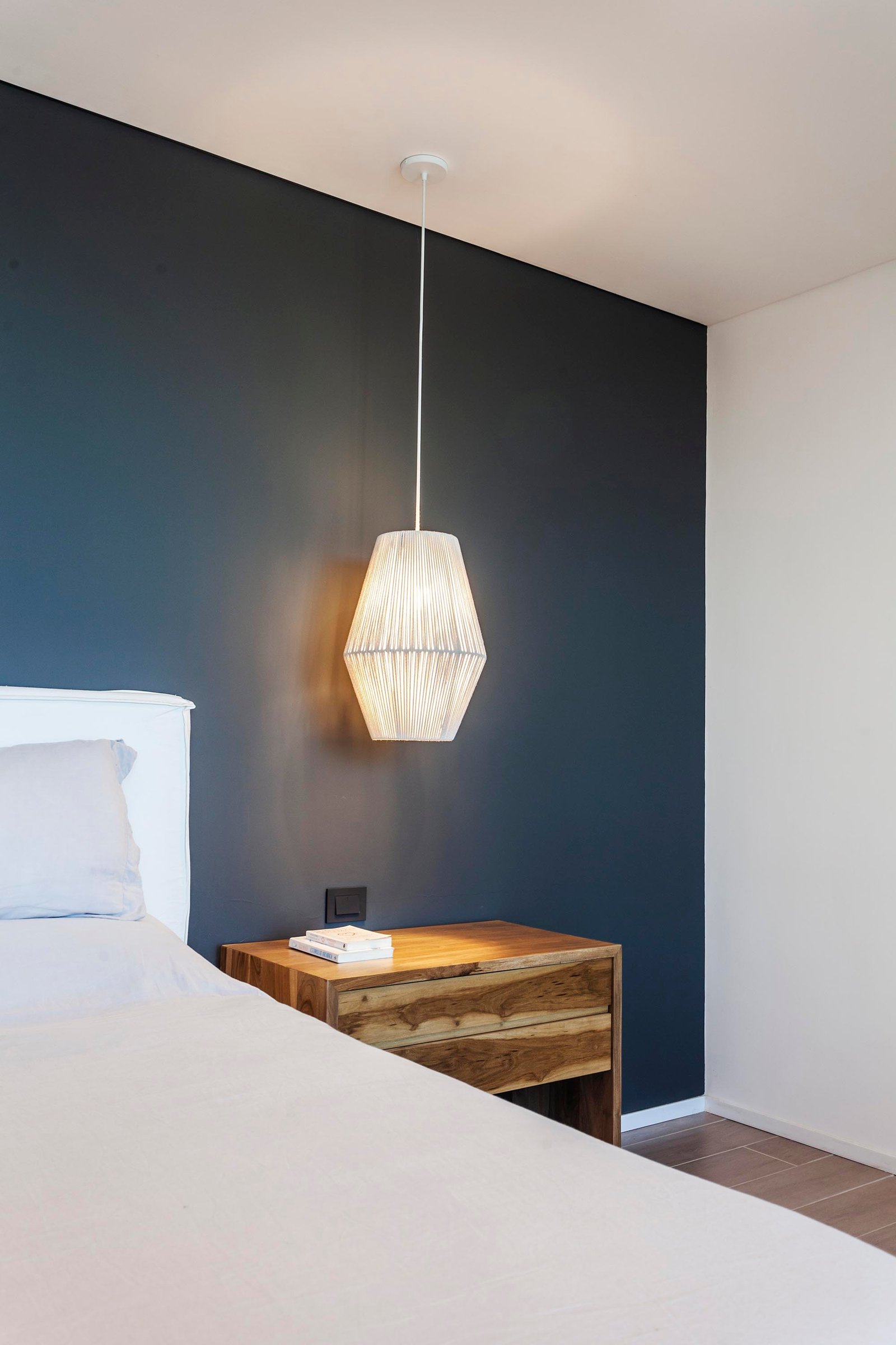 Dormitorio moderno con pared azul oscuro, lámpara colgante tejida y mesita de noche de madera — proyecto CASA C313, por A'PRIMA - Arquitectura Sustentable, en Provincia de Buenos A