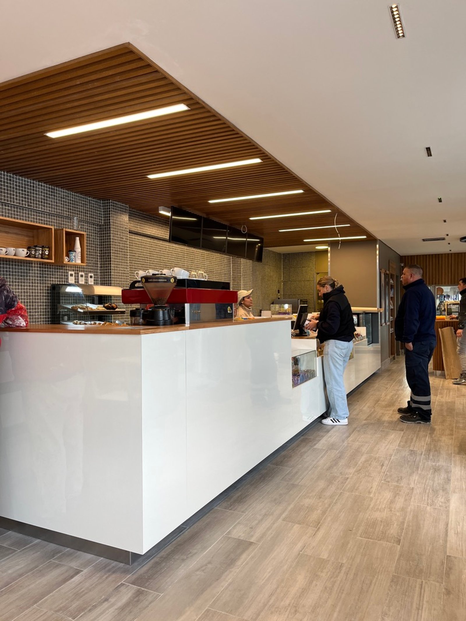 Interior de cafeteria moderna com teto de ripas de madeira e balcão branco — projeto Full YPF Muniz, por Arcucci Arquitectura, em Muñiz