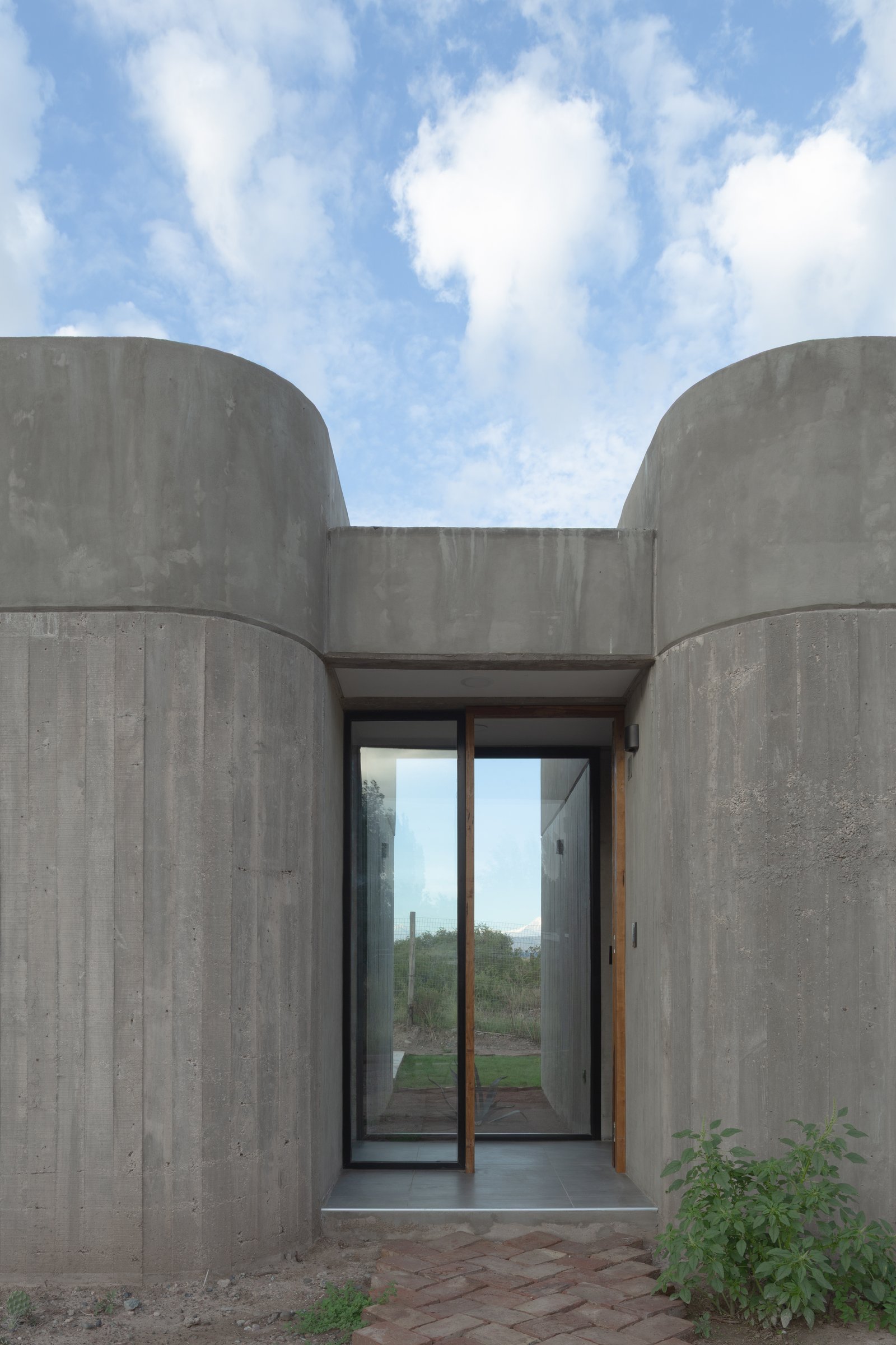 fachada de concreto aparente com entrada de vidro e paredes curvas — projeto Complejo SCV, por ONA - Oficina Nómada de Arquitectura, em Las Compuertas