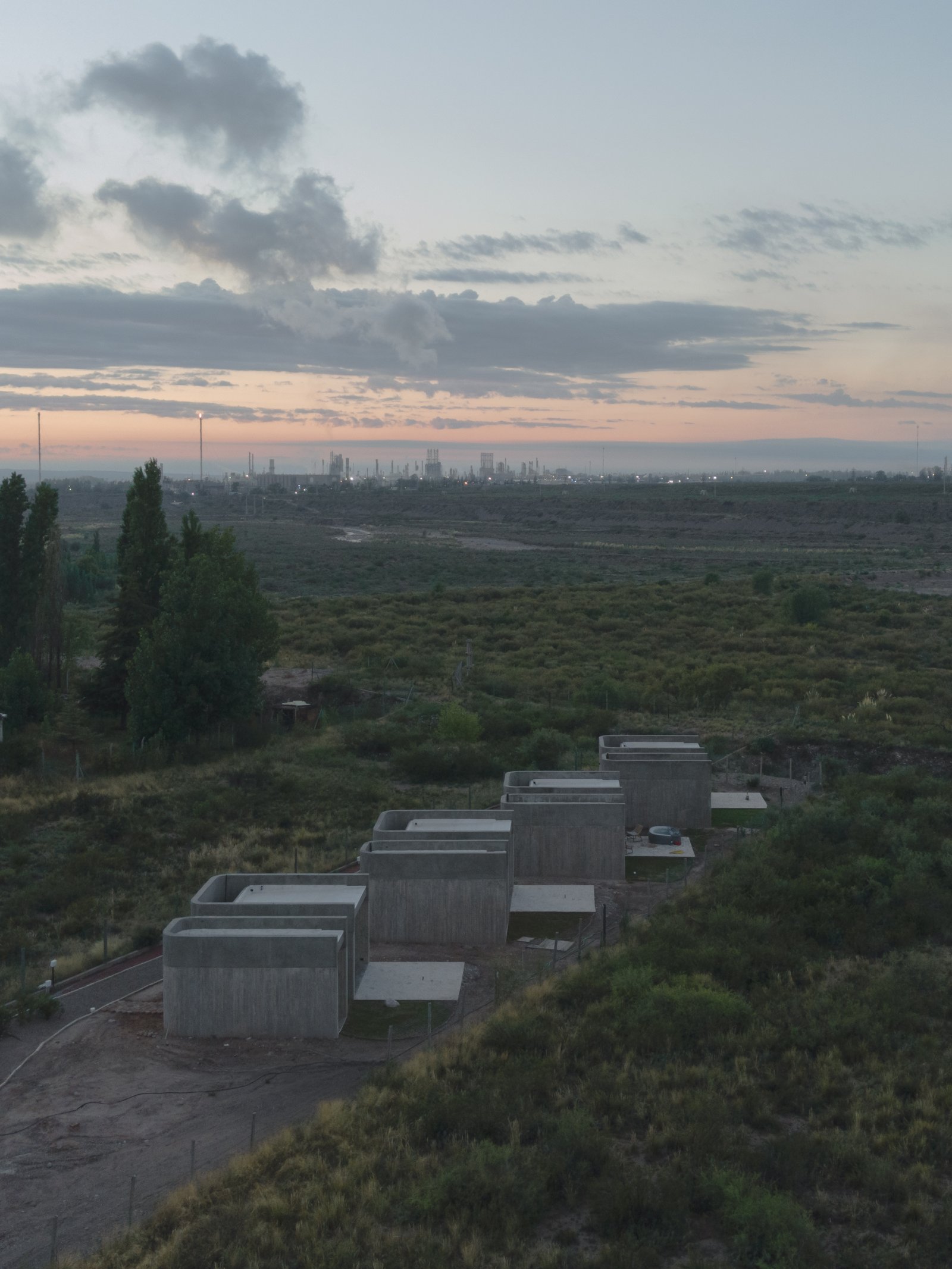 edifício moderno de concreto em colina rural com indústria ao fundo — projeto Complejo SCV, por ONA - Oficina Nómada de Arquitectura, em Las Compuertas