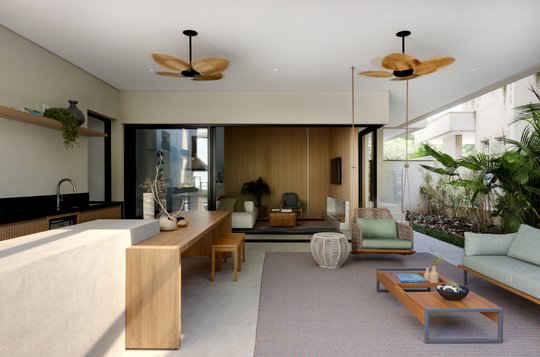 Amplio living comedor con cocina integrada, muebles de madera clara y ventiladores colgantes — proyecto Projeto Destaque Club&Casa, por Barbara Dundes – Arquitetura + Design