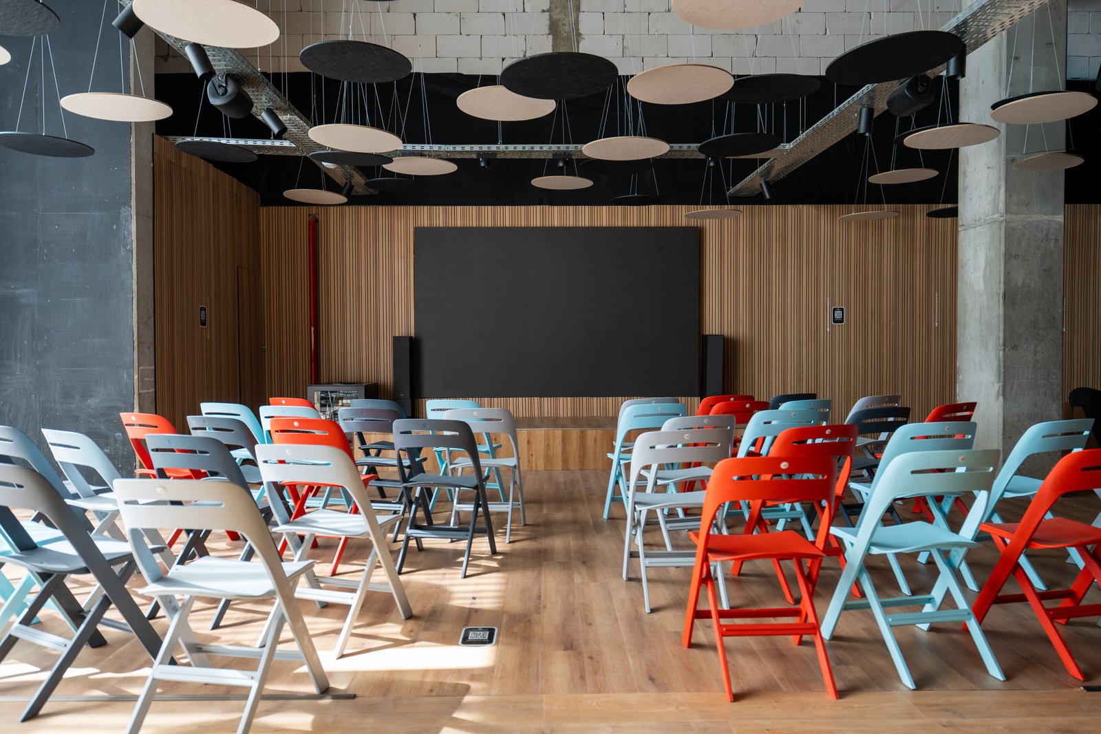 Sala de conferências com cadeiras azuis e vermelhas e paredes de madeira — projeto Auditorio LN, por KUPERDESIGN CONSTRUCCIONES SRL, em Buenos Aires