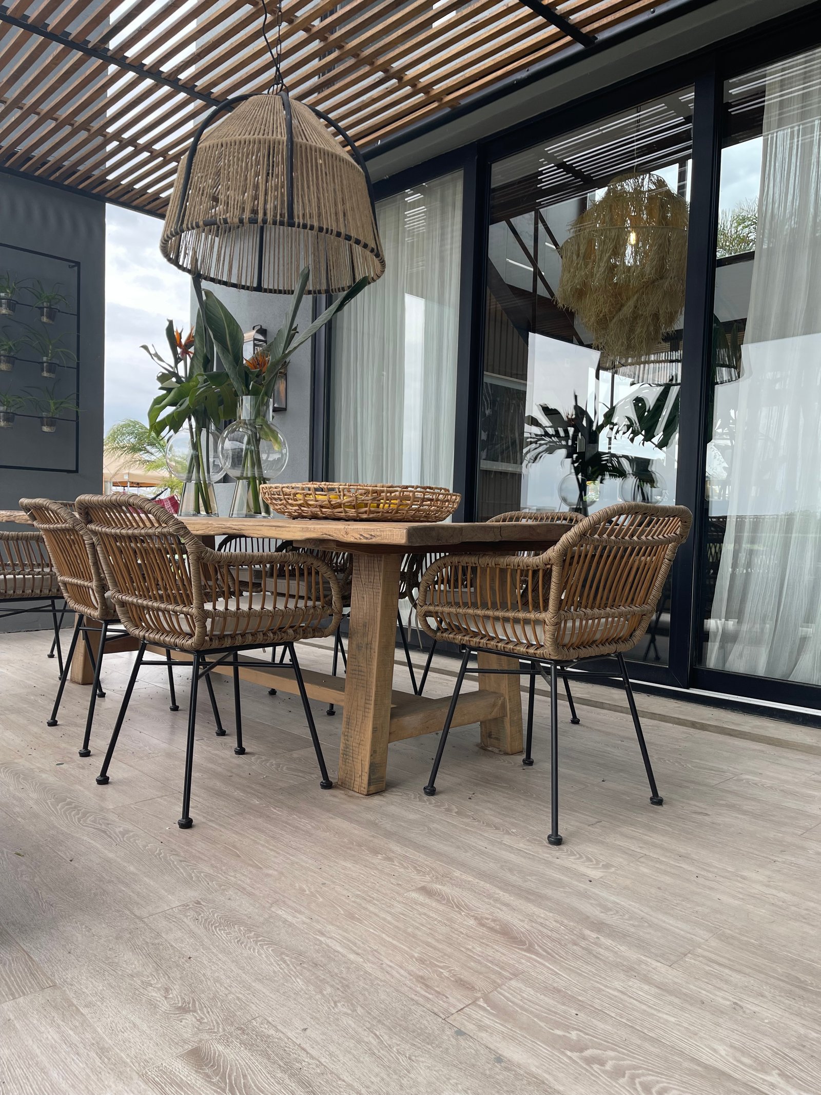 Comedor exterior con sillas de ratán y mesa de madera bajo pérgola — proyecto Maderas, por Vite Porcellanato