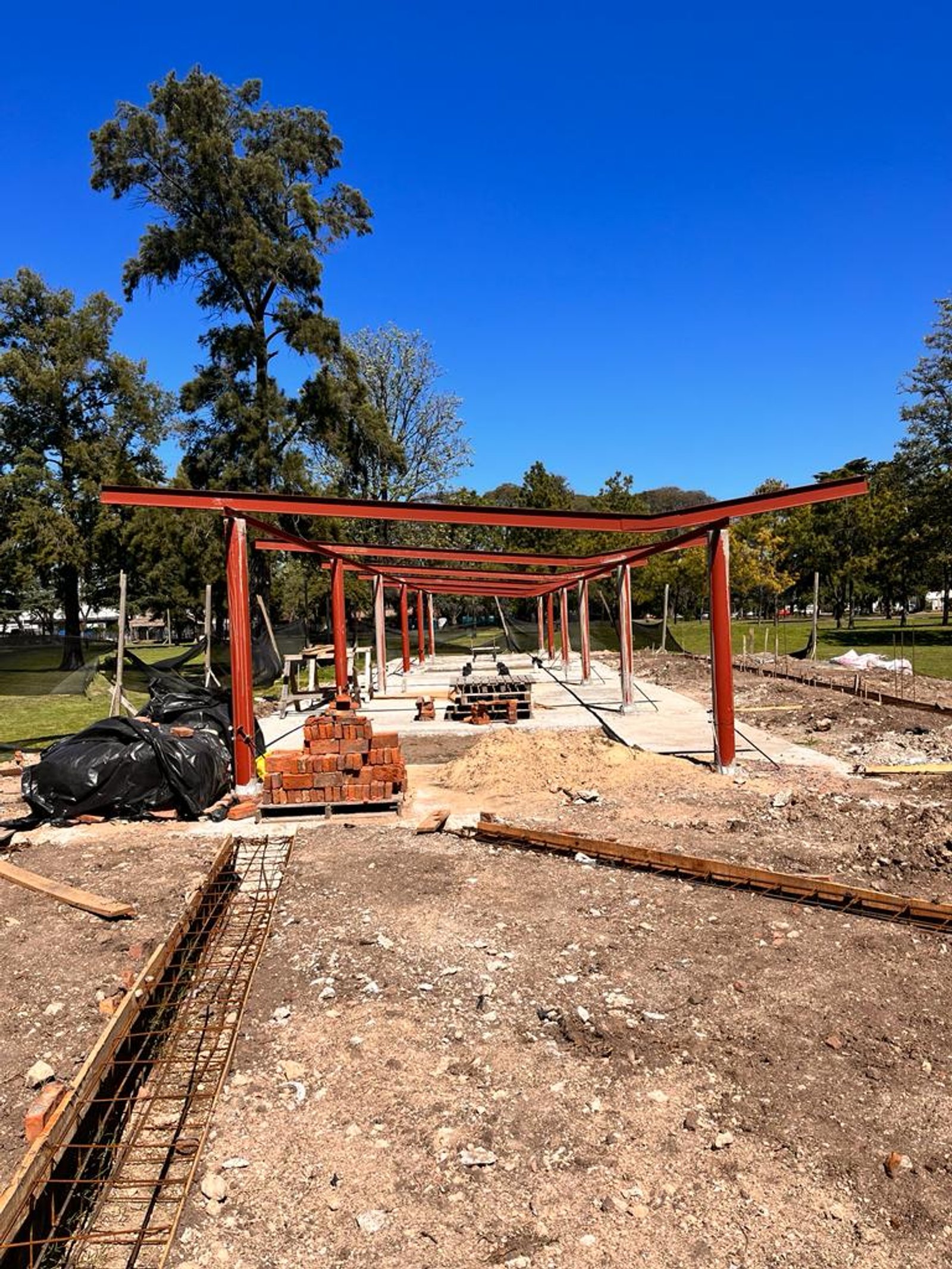 Obra de pérgola roja de metal, cielo azul, estructura en progreso — proyecto Parque Público Villa Adelina, por Estudio Rifahi, en B1607 Villa Adelina