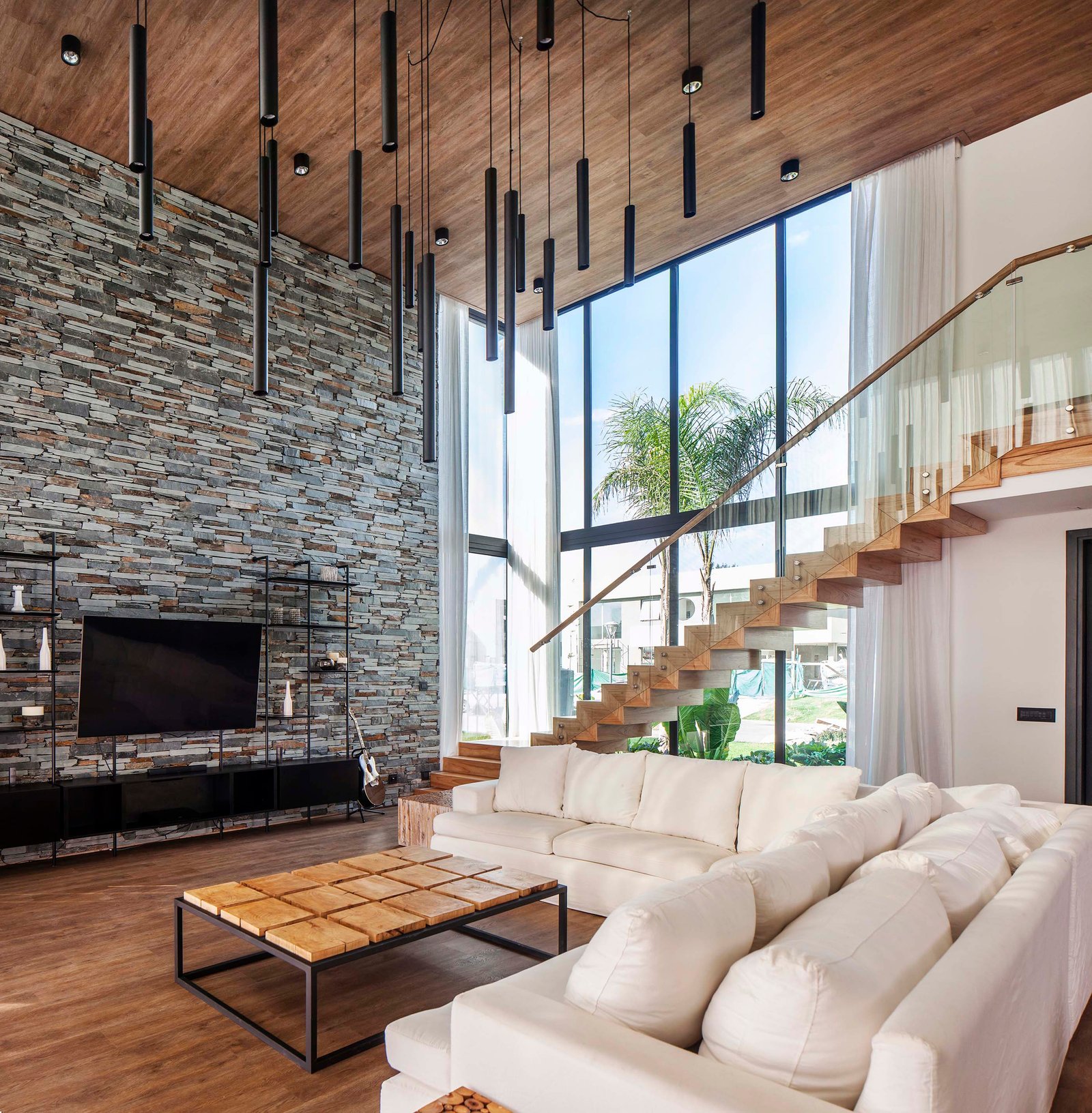 Living moderno de doble altura con pared de piedra y escalera flotante — proyecto CASA T199, por A'PRIMA - Arquitectura Sustentable, en Los Robles 5901