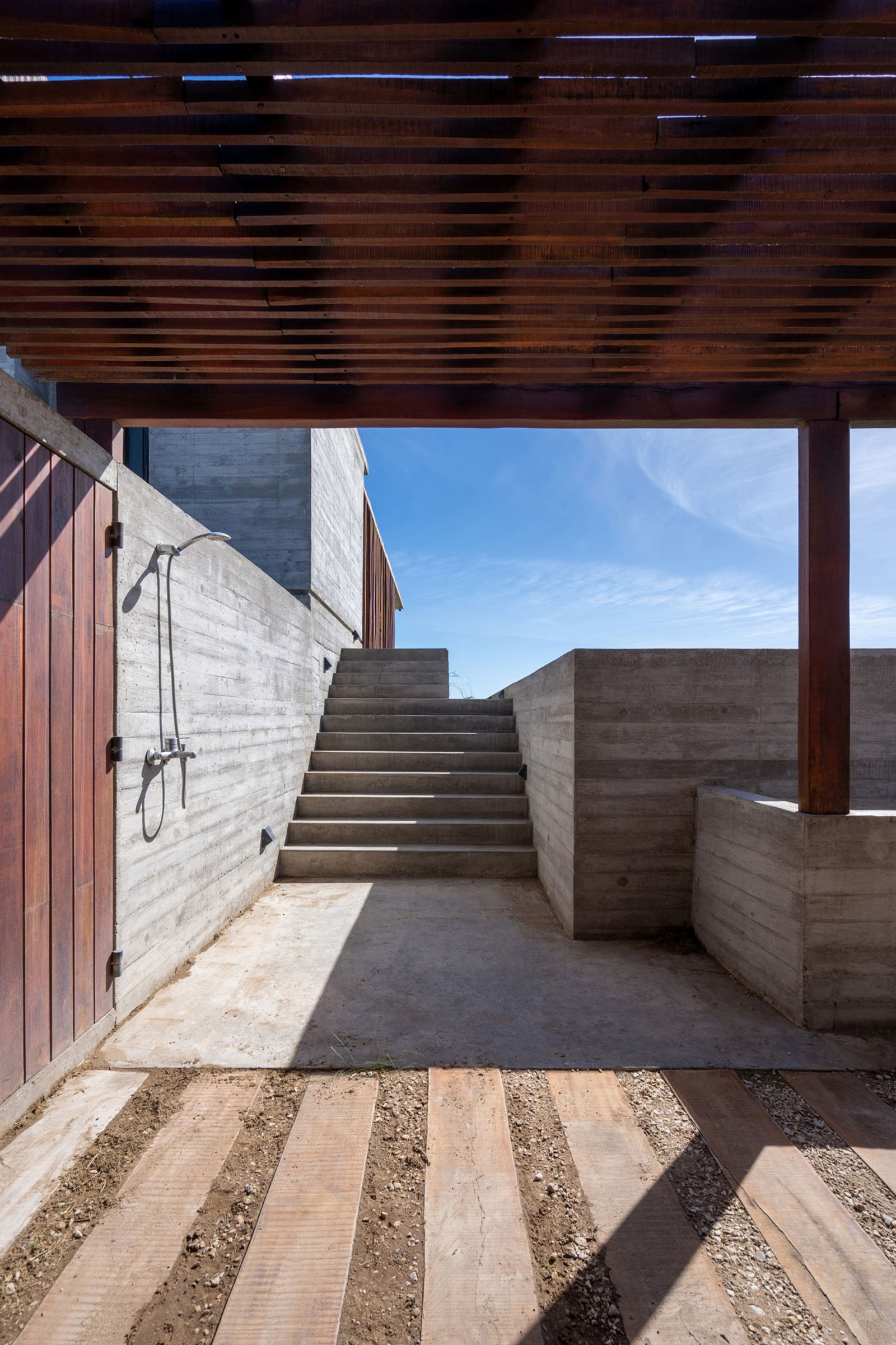 espaço externo brutalista com chuveiro, pergolado de madeira e escada — projeto Casa Médanos, por Besonías Almeida arquitectos, em JWRC+MQ