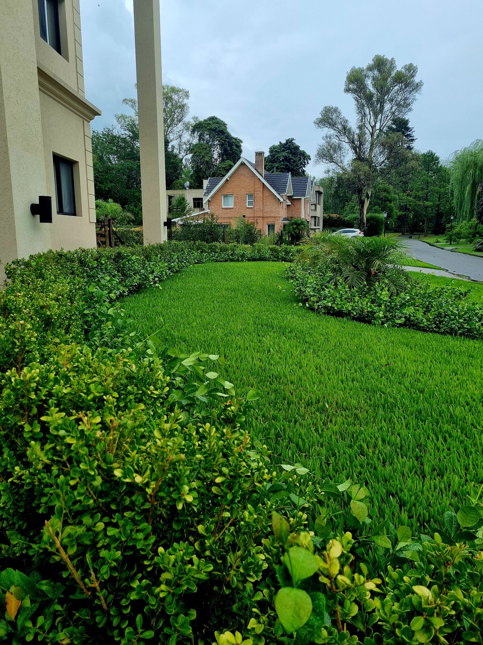 Fachada de casa bege com arbustos, gramado e casa de tijolos — projeto Abril Club de Campo, por Marcela García Brünner Paisajista, em B1885 Guillermo Enrique Hudson