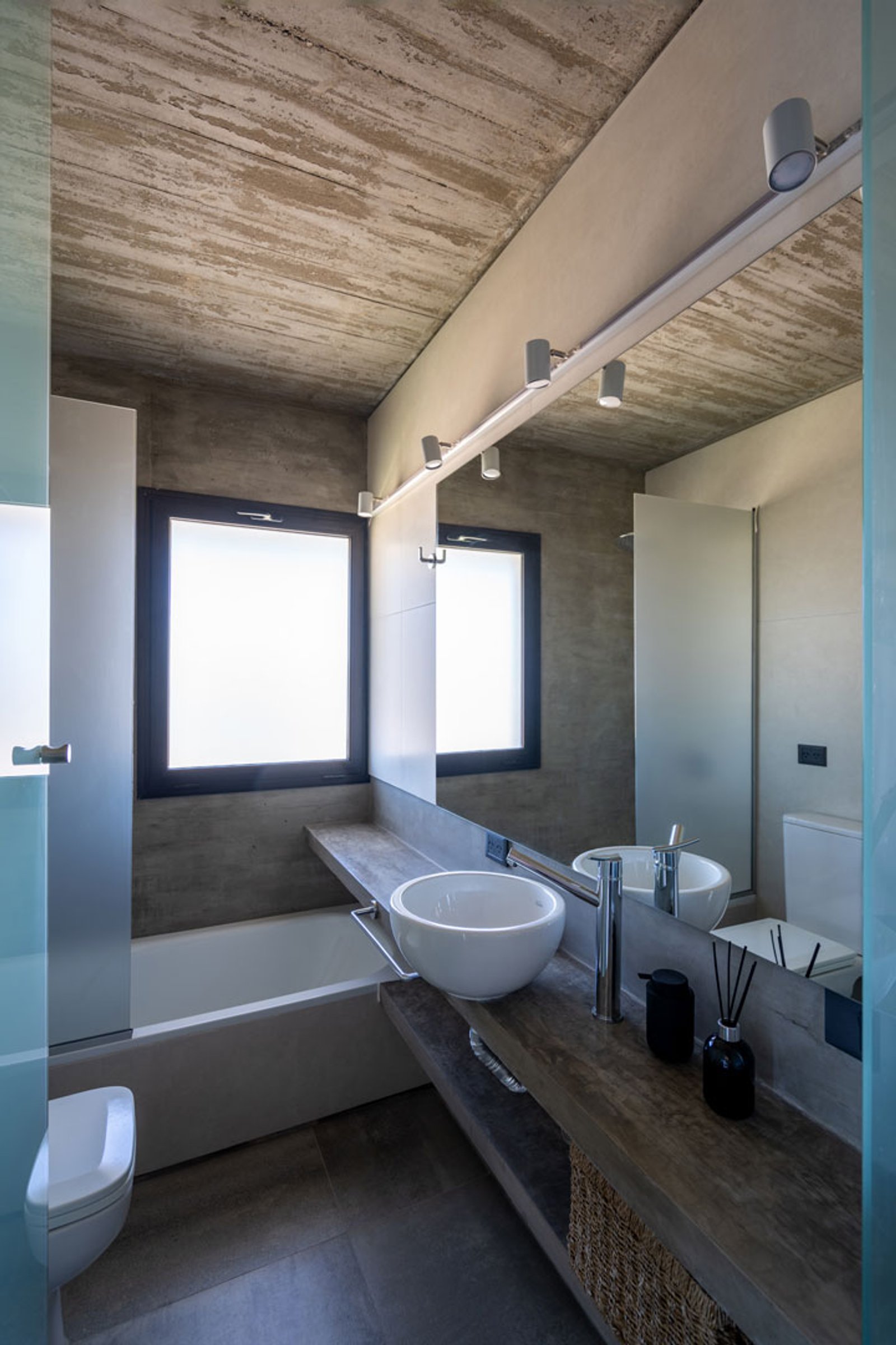 Baño moderno industrial con hormigón expuesto, bacha de cerámica y luces integradas — proyecto Casa La Marina, por Besonías Almeida arquitectos, en X6VF+67