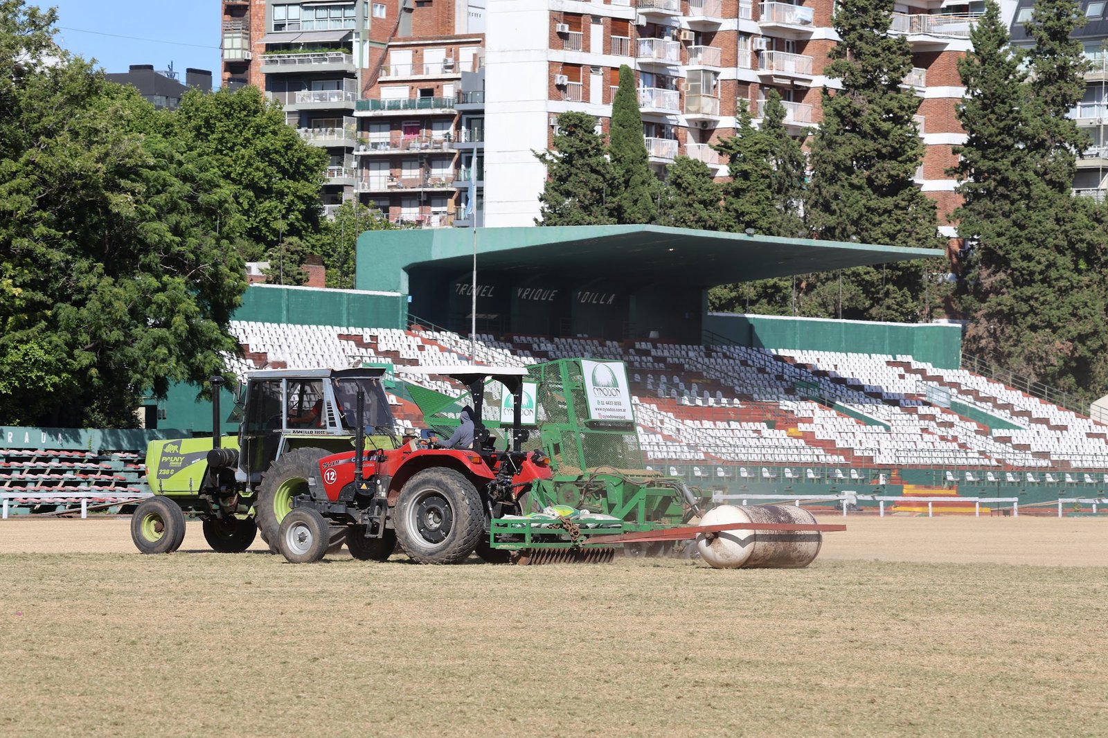 Trator vermelho em campo com arquibancadas de estádio e prédio — projeto Row planting Palermo Polo, por Cynodon, em Av. del Libertador 4096