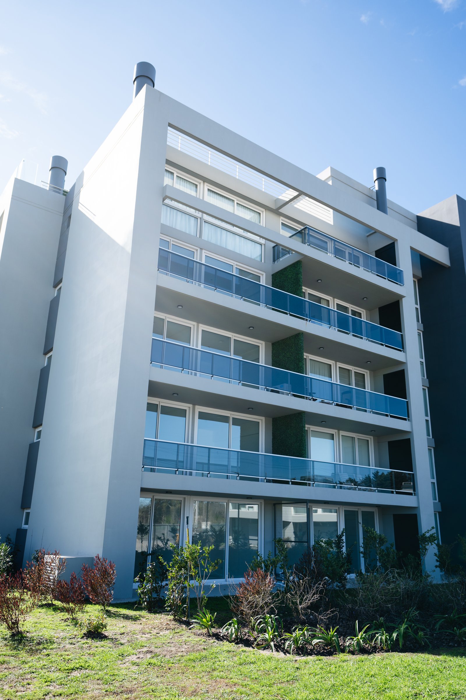Edificio moderno de varios pisos con fachada blanca y balcones de vidrio azul — proyecto Luciérnagas, por MUCHTEK, en Pilar
