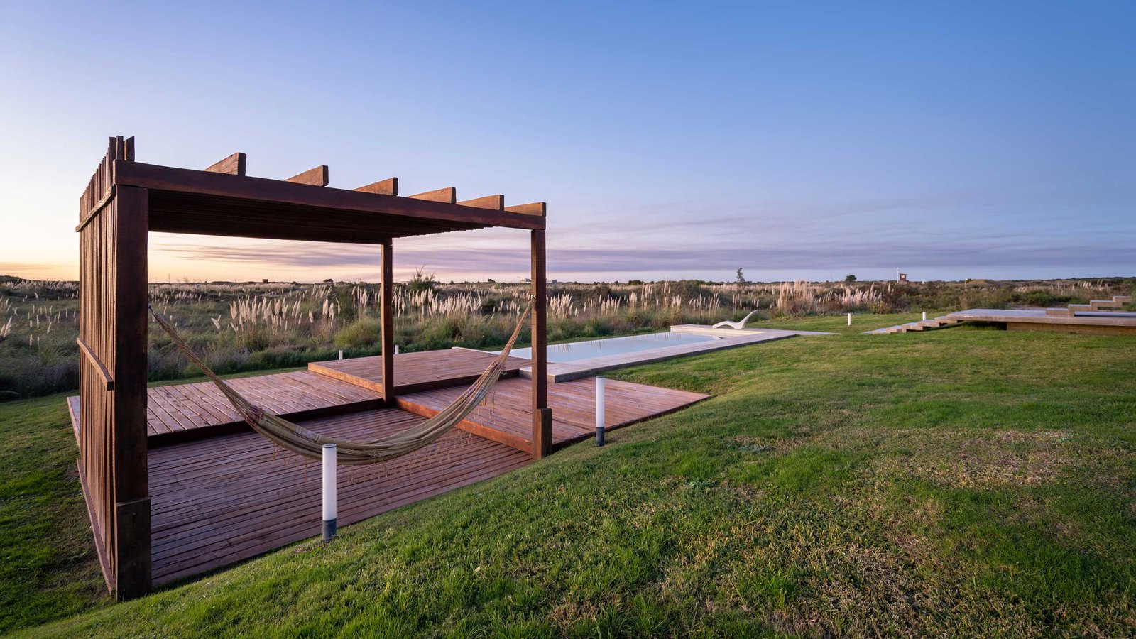 Área de piscina externa com pergolado, deck de madeira e rede — projeto Casa Médanos, por Besonías Almeida arquitectos, em JWRC+MQ