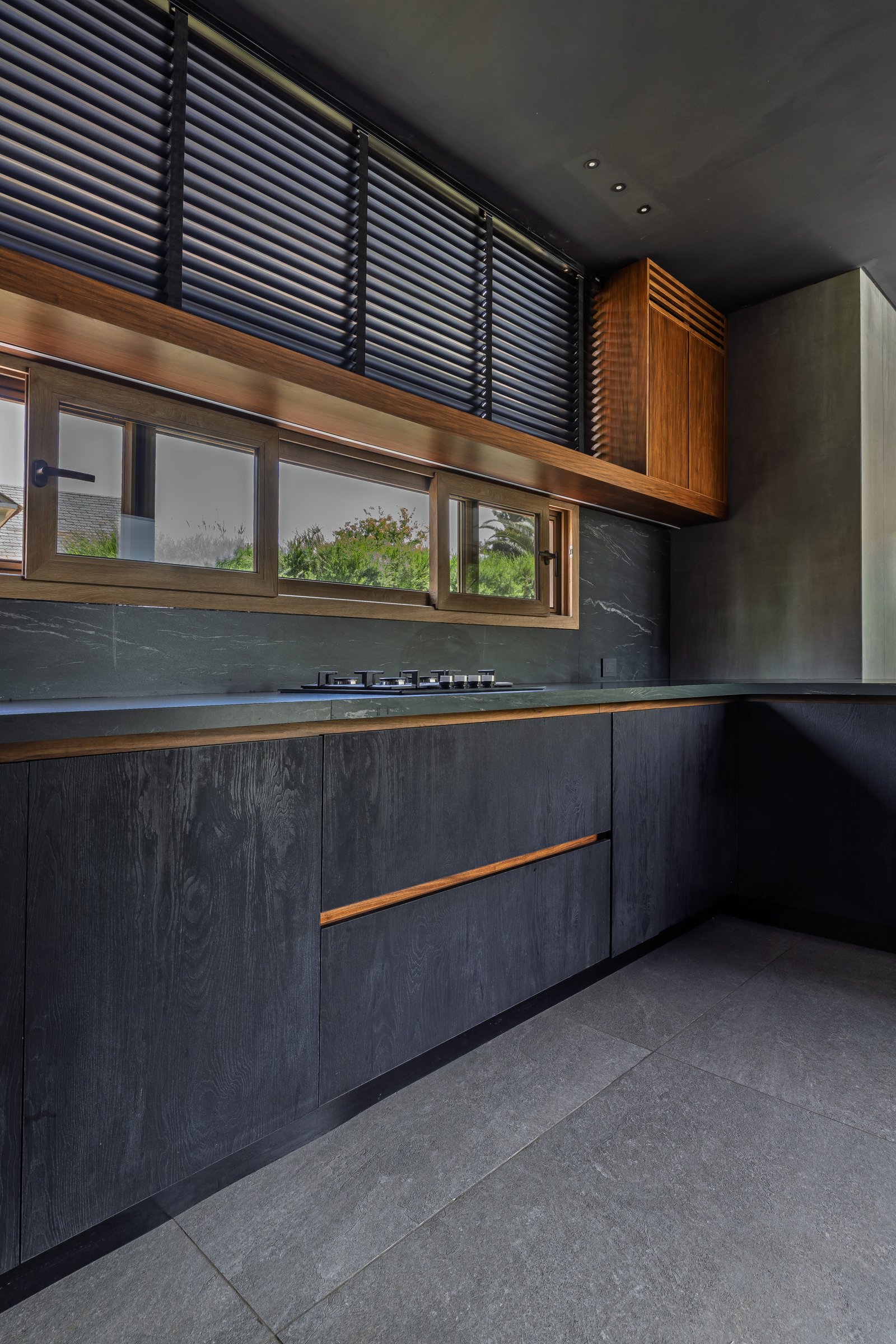 Cocina moderna con gabinetes oscuros, mesada de piedra y ventana grande — proyecto Castores - Estudio Gambotto, por ABEJA CONCEPT, en Los Castores