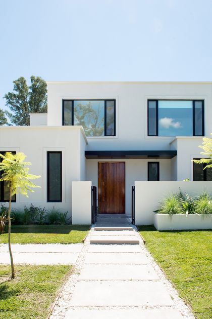 Moderna fachada blanca con grandes ventanales y camino de losas — proyecto Bosque Chico, por Estudio Arq. María Sioli, en Garin