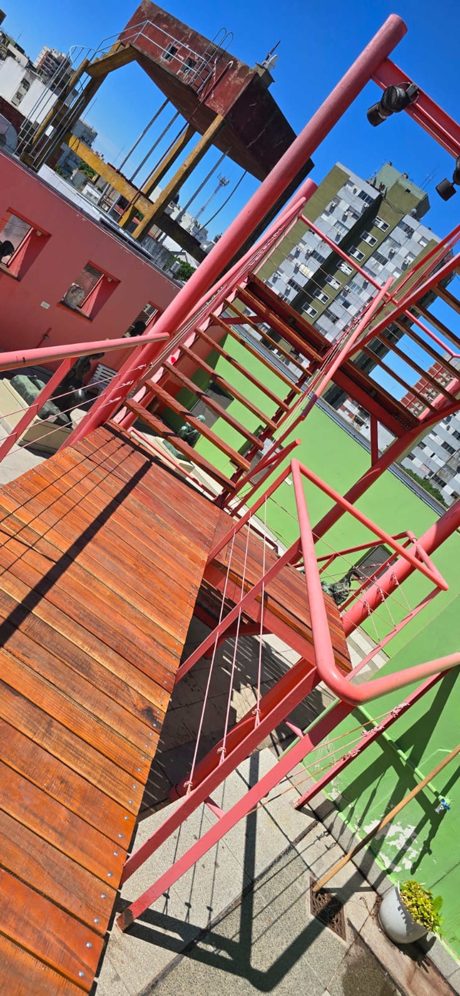 Escalera metálica roja sobre deck de madera con fachada rosa y pared verde en entorno urbano — proyecto Museo Benito Quinquela Martin, por ASV Muebles y Maderas, en Av. Don Pedro d