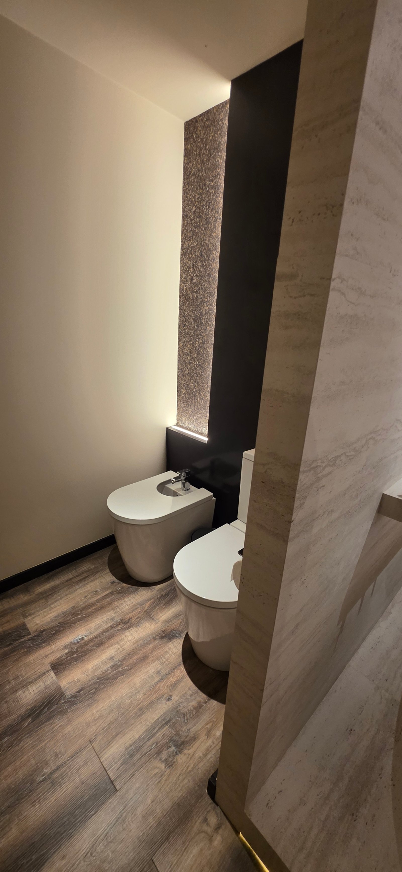 Baño moderno con dos inodoros, suelo de madera oscura y luz indirecta — proyecto Baño en suite sencualidad natural UNE 2026 Posadas Misiones, por Matec ss, en Posadas