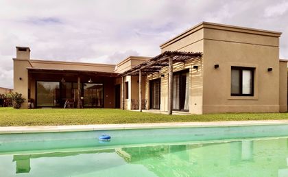 Casa moderna con fachada beige, galería de madera y pileta verde — proyecto Santa Teresa 2, por Marconi Silva Arquitectos, en Benavidez