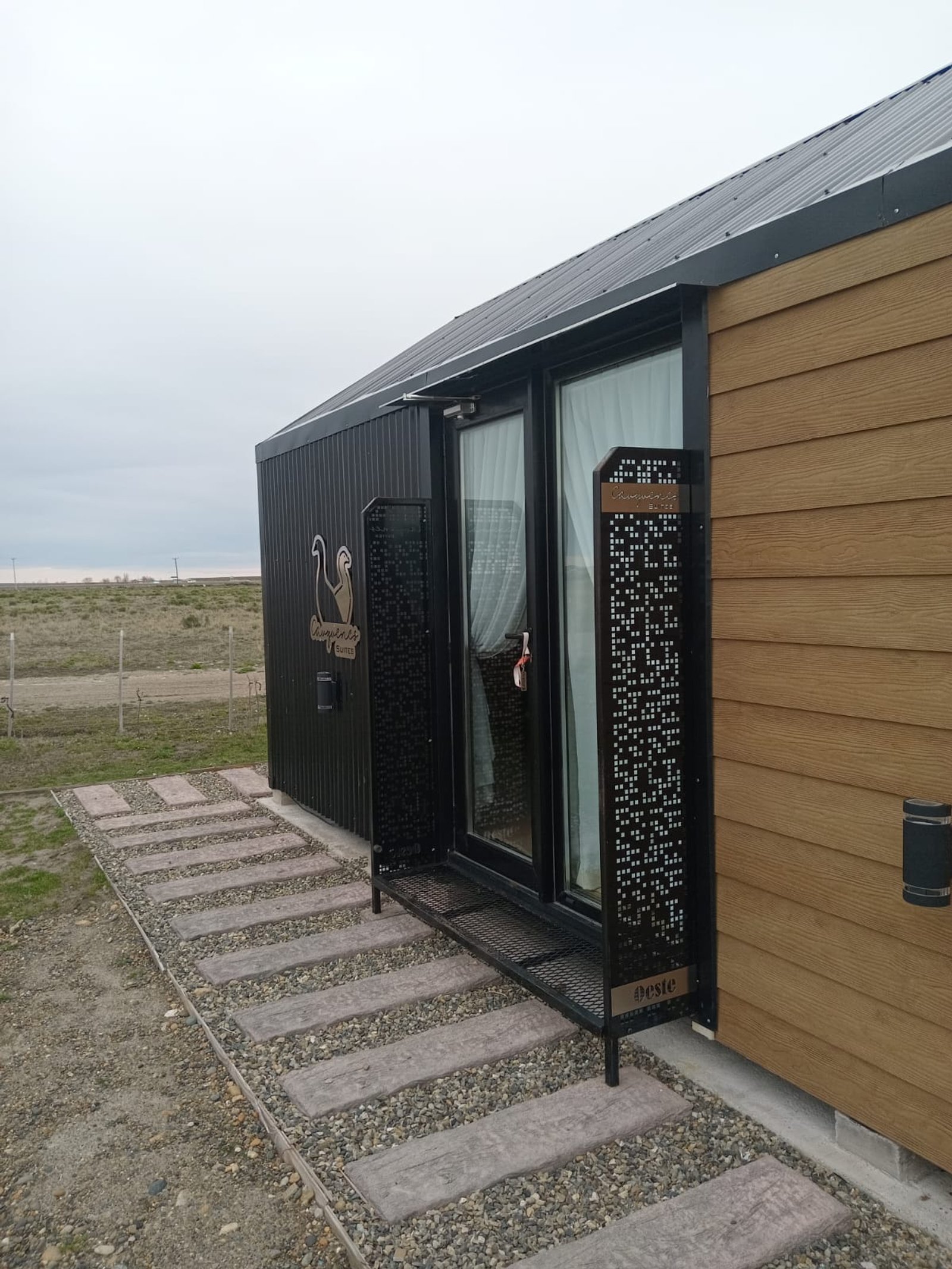 Fachada de casa moderna con revestimiento de madera y metal, puerta de cristal y camino de piedra — proyecto Rio Gallegos Tiny Houses, por TROKEN, en Río Gallegos