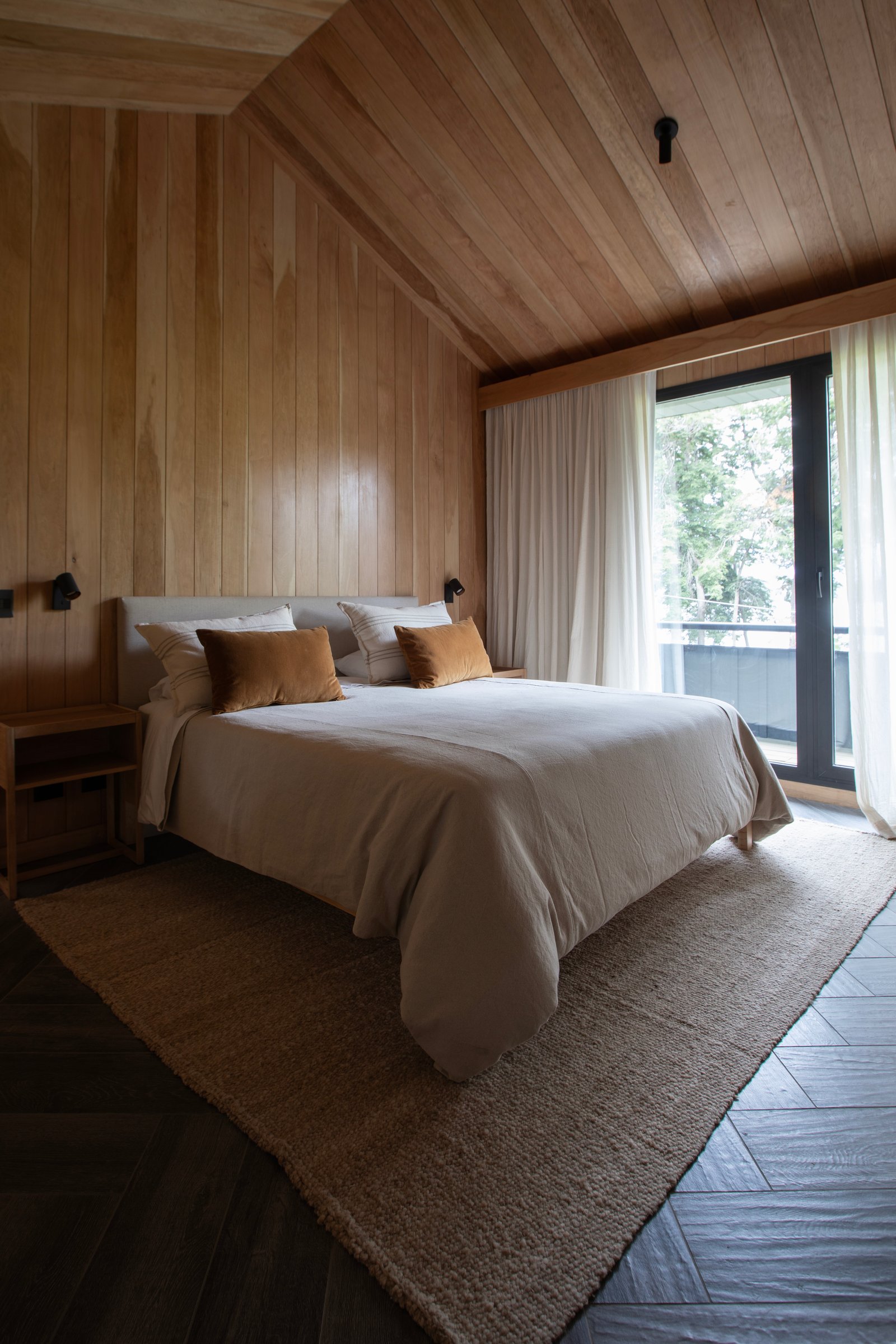 Quarto rústico moderno com paredes e teto de madeira, cama bege e detalhes em laranja — projeto OBRA SANTA BARBARA, por ILVA, em Provincia de Buenos Aires