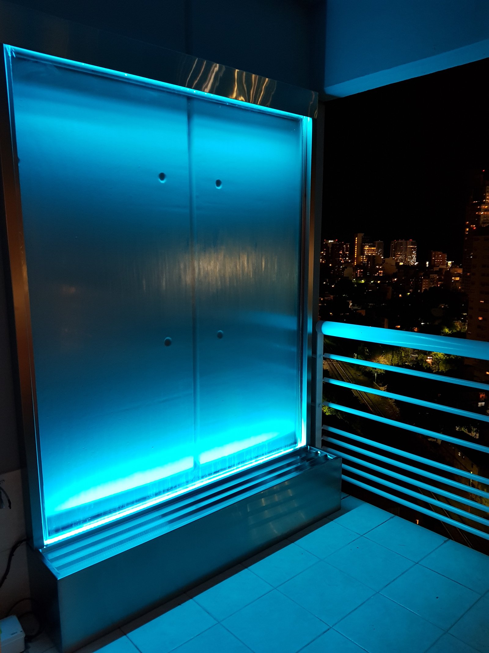 Terraza moderna con cascada de agua iluminada y vista urbana nocturna — proyecto Fuente Agua Balcon , por FUENTES Y MUROS .COM.AR, en B1636 Olivos