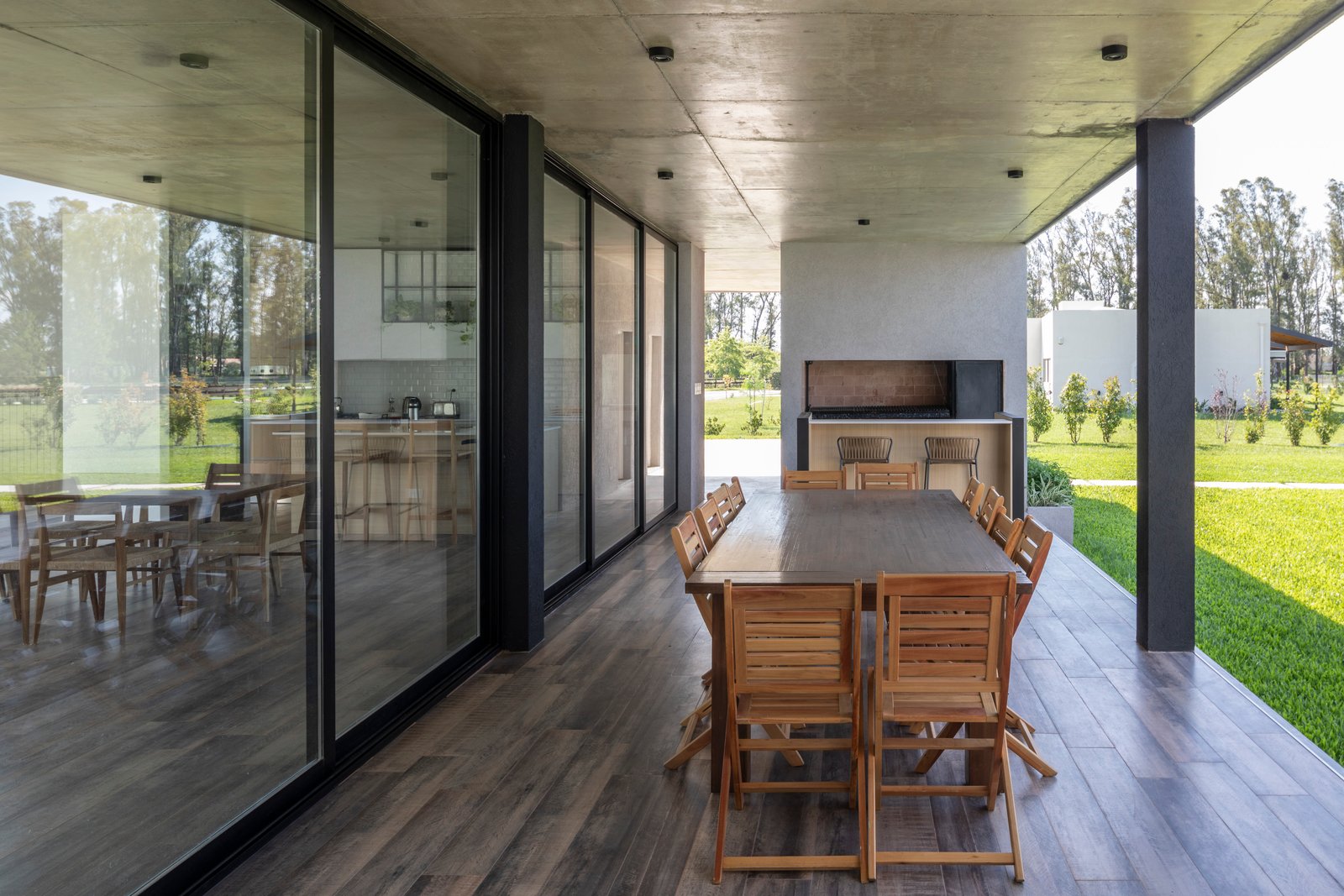 Área de jantar externa moderna com deck de madeira e churrasqueira, ao lado de grandes janelas — projeto Casa Cruz, por Grupo CRBN, em B2812 Exaltación de la Cruz