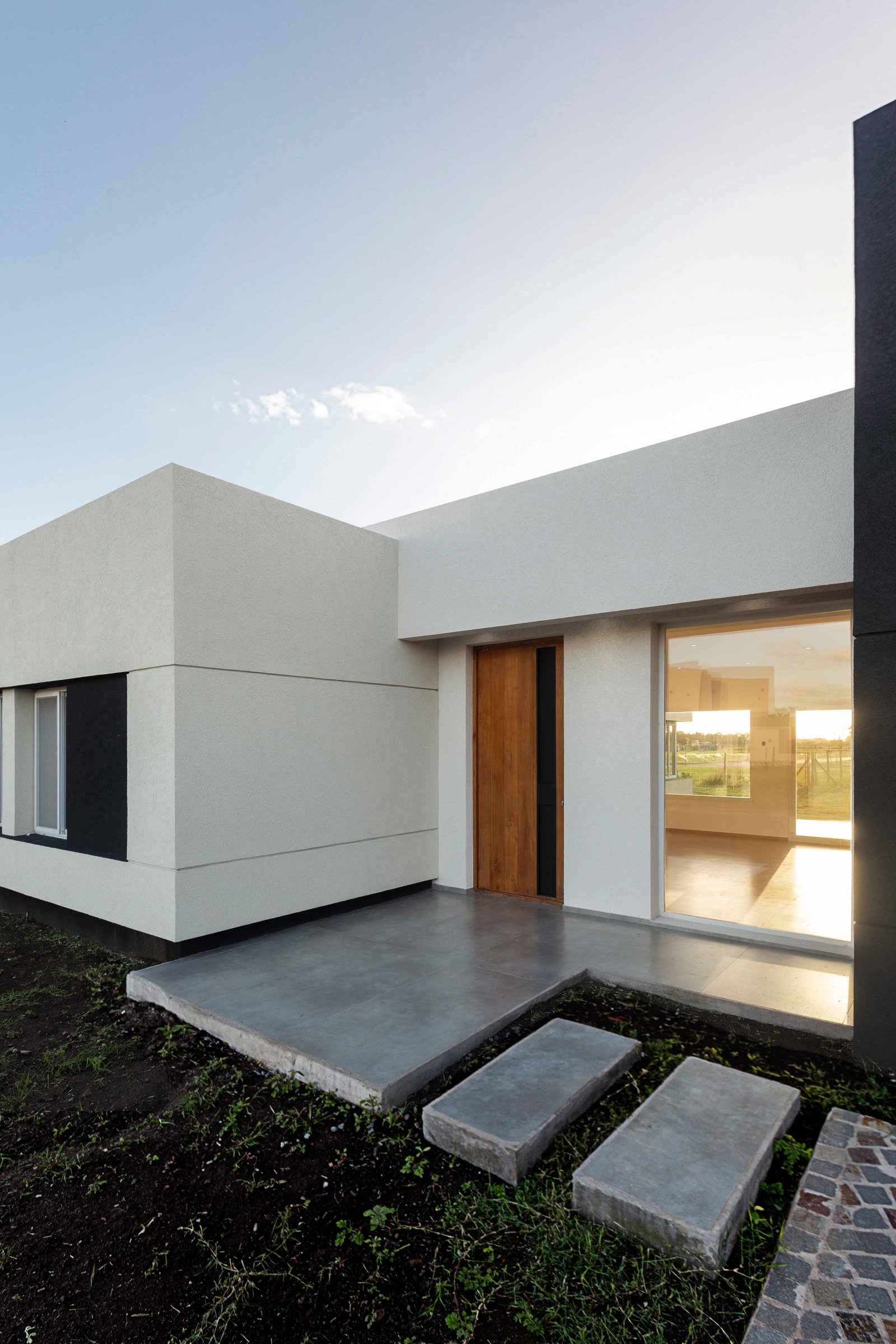 Fachada moderna branca com porta de madeira e janela do chão ao teto — projeto San Simon I, por Estudio Stepa, em Ezeiza