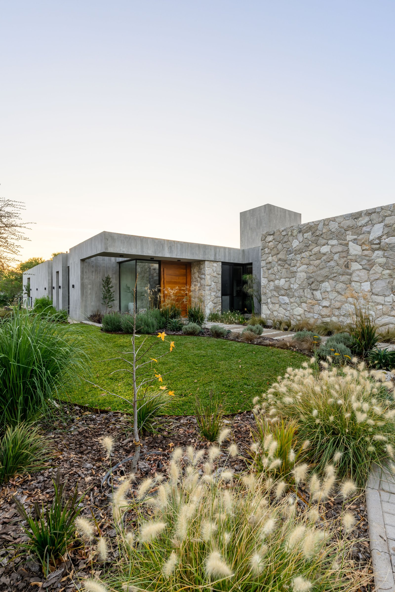 Fachada moderna de piedra y hormigón con jardín exuberante — proyecto La Paloma :: Villa Allende - Córdoba :: Paisajismo Residencial :: STIPA Arte Nat, por STIPA Arte Natural, en M