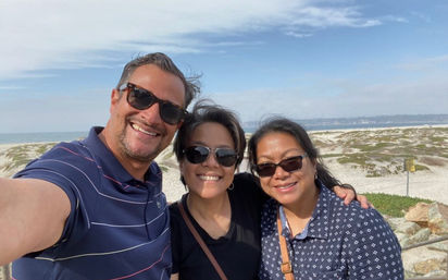 Three smiling adults wearing sunglasses pose for a seaside selfie with sandy dunes, rocks and the ocean under a partly cloudy sky.