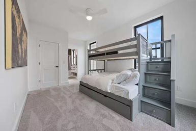 Bright modern bedroom interior with gray wooden bunk bed, lower trundle and built-in staircase drawers, white bedding, plush gray carpet, white walls and windows flooding the room with natural light.