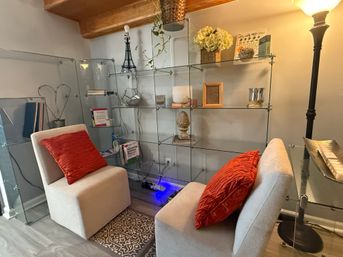 Cozy modern reading nook with two beige armless chairs topped with red textured pillows, glass modular shelving filled with books and decorative accents, a tall floor lamp, patterned rug and warm wooden ceiling beam.