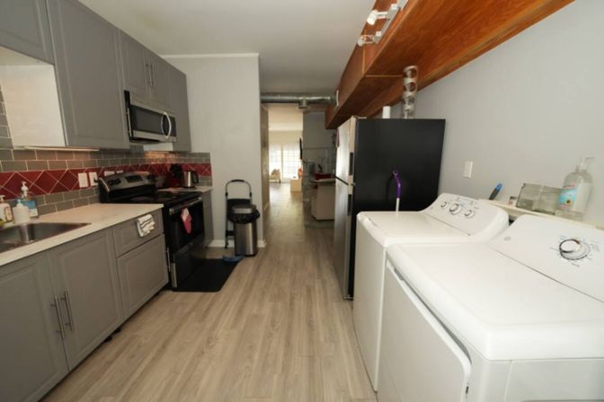 Galley apartment kitchen with side-by-side washer and dryer, gray cabinets, stainless stove and refrigerator, red tile backsplash and light wood laminate floor
