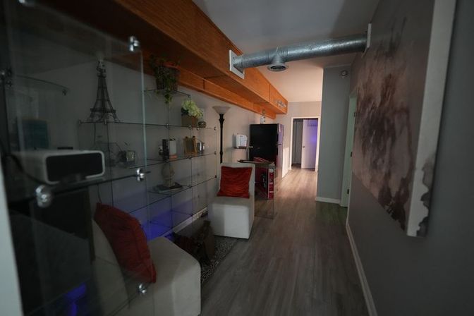 Modern urban loft hallway with exposed metal duct and wooden beam, glass display shelves with decor, white chairs with red pillows, gray wood-look flooring and a kitchenette visible at the far end.