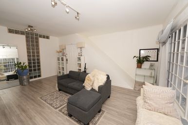 Bright modern living room with gray sectional sofa and ottoman on light wood floors, glass-block accent wall, large grid windows, neutral pillows and faux-fur throw, area rugs, plants and minimalist shelving.