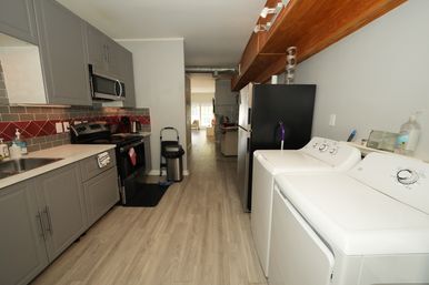 Modern galley apartment kitchen with gray cabinets, stainless appliances, washer and dryer, wood-look flooring and open-plan hallway to a bright living area.