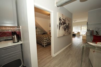 Modern apartment hallway with light wood floors, kitchenette and bedroom doorway to the left with a patterned bedspread, abstract wall art, exposed ceiling duct, and a bright open living room with large windows.