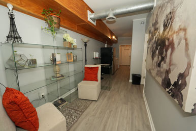 Loft-style apartment hallway with glass display shelves, beige chairs with red throw pillows, exposed metal ductwork under a wood beam, large abstract wall art, light wood floors and a TV near the entry.