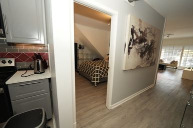 Sunlit modern apartment hallway with wood-look floors, grey kitchen cabinets and kettle, open doorway to a bedroom with patterned duvet, abstract wall art and bright living room with large windows.