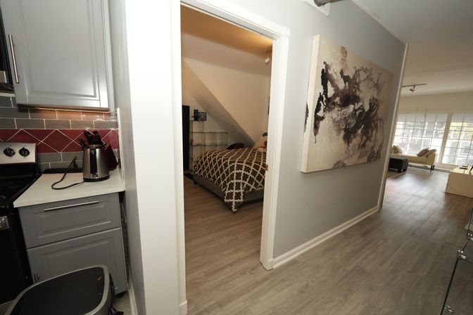 Sunlit modern apartment hallway with wood-look floors, grey kitchen cabinets and kettle, open doorway to a bedroom with patterned duvet, abstract wall art and bright living room with large windows.