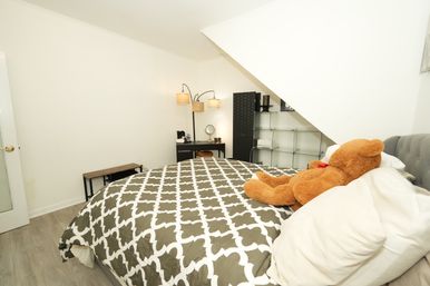 Cozy attic-style bedroom with sloped ceiling, gray-and-white patterned duvet, large brown teddy bear on pillows, small desk with mirror and floor lamp, glass shelving and wood floors.