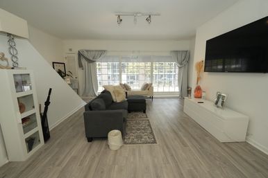 Bright, airy modern apartment living room with a gray sectional and chaise on light wood floors, large paned windows with soft drapes, white media console and wall-mounted TV, neutral pillows and area rugs.