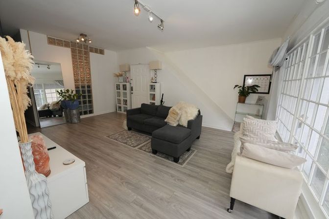 Bright modern living room with light gray wood-look floors, large grid windows, dark gray sectional with chaise on area rugs, cream bench with throw pillows, glass-block accent wall, potted plants and minimalist decor.