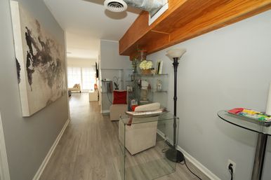 Inviting modern apartment hallway with light gray walls and wood-look floors, a large abstract canvas on the left, glass shelves and beige chairs with red pillows beneath an exposed wooden beam and tall floor lamp, leading to a bright open living room.