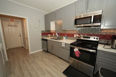 Small modern kitchen with gray cabinets, stainless-steel appliances, red diamond tile backsplash, sink and dishwasher, light wood laminate floor and hallway with EXIT sign.